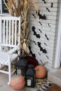 A picture of white rocking chair on a front porch surrounded by hanging bats and pumpkins.
